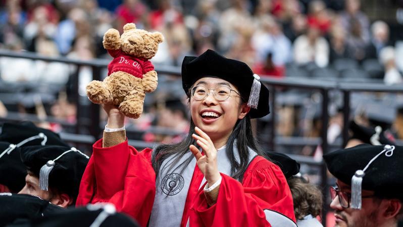 Ohio State doctoral graduate in academic regalia holding up a teddy bear during the 2023 autumn commencement ceremony