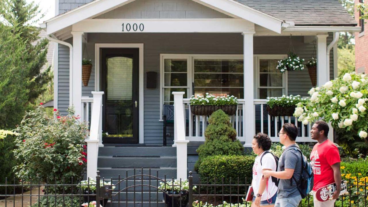 Three students walking on a sidewalk in front of a gray house in the spring